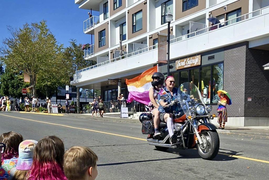 Two people ride down the street on a motorbike with a large lesbian flag waving behind them