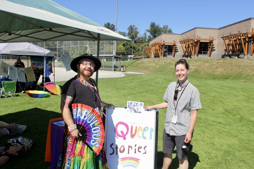 Two people stand in front of a sign that says 'Queer stories.' One holds a fan that says 'fabulous'
