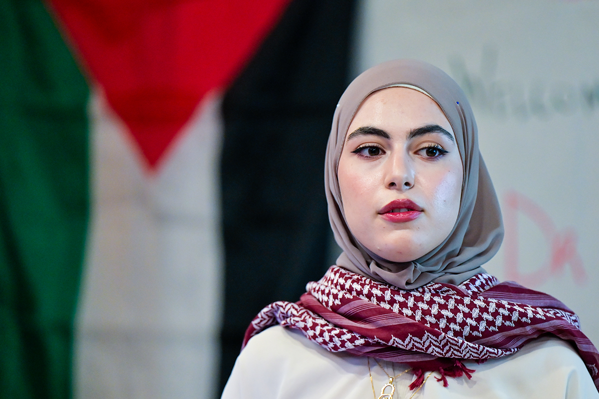 A woman wearing a headscarf and keffiyeh stands in front of the Palestine flag.