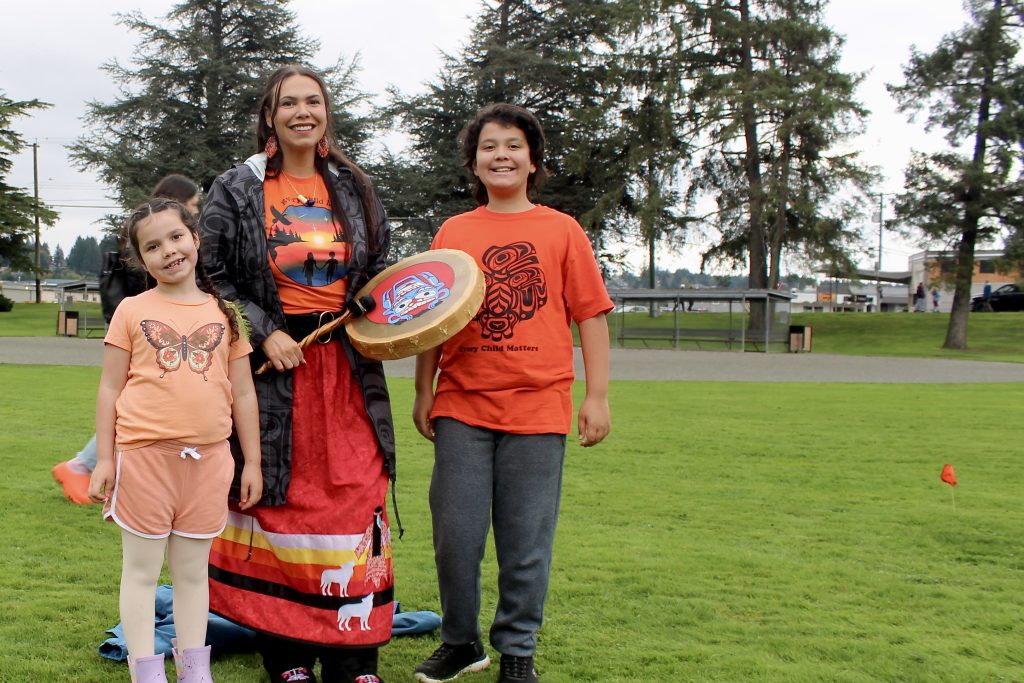 An Indigenous woman with an orange shirt and ribbon skirt holds a drum with Coast Salish art on it. She is standing in between her two children, who also both have orange shirts. All are smiling.