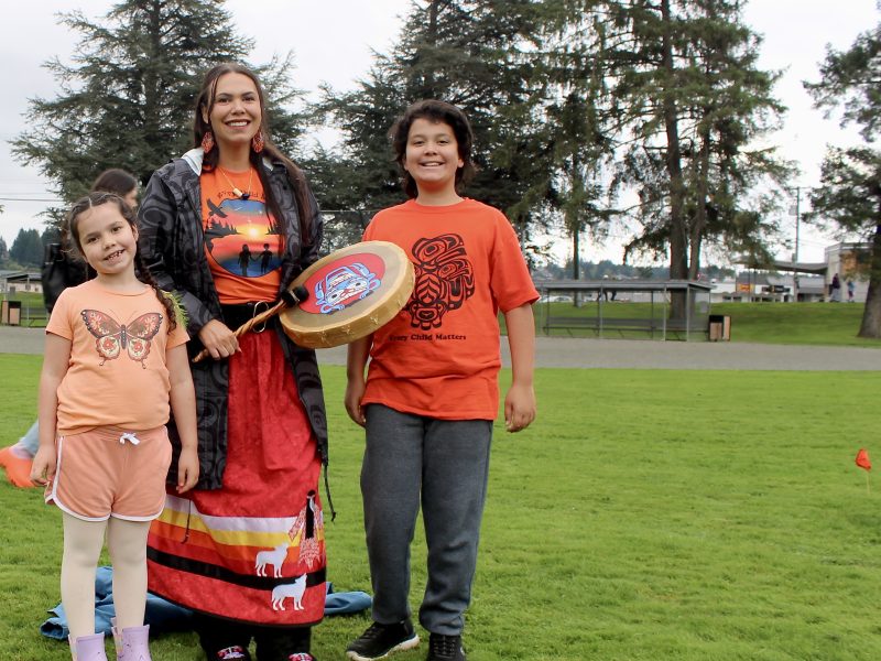 An Indigenous woman with an orange shirt and ribbon skirt holds a drum with Coast Salish art on it. She is standing in between her two children, who also both have orange shirts. All are smiling.
