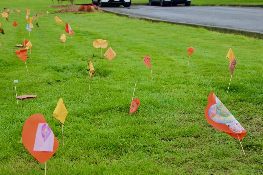 Orange paper hearts on sticks are arranged in the grass. They have different poems or drawings on them.