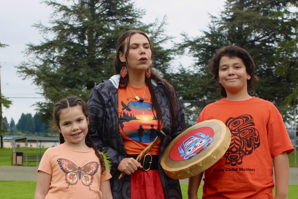 An Indigenous woman with an orange shirt and ribbon skirt plays a drum with Coast Salish art on it. She is standing in between her two children, who also both have orange shirts.