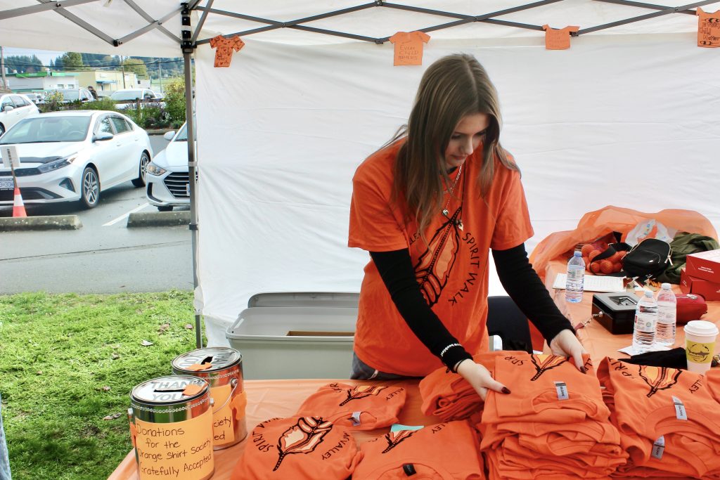 A woman with long brown hair in an orange shirt stands at a volunteer booth and folds a pile of orange shirts. There are two donation cans on the table next to the shirts.