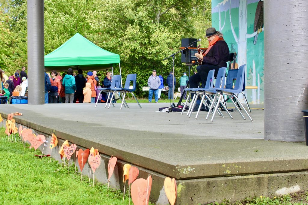 A man in a hat plays guitar on an outside stage. On the grass below the front of the stage there is a line of orange paper hearts. 