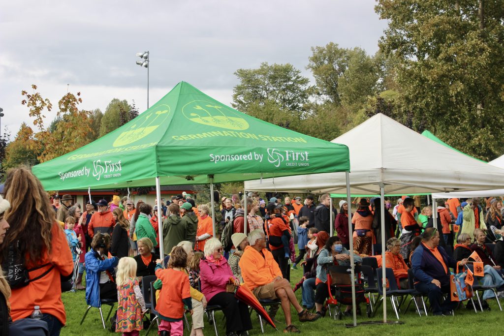 A crowd of people in orange shirts gather under volunteer tents on a rainy day. Some sit in lawn chairs and appear to be watching something, and some stand and chat with one another.