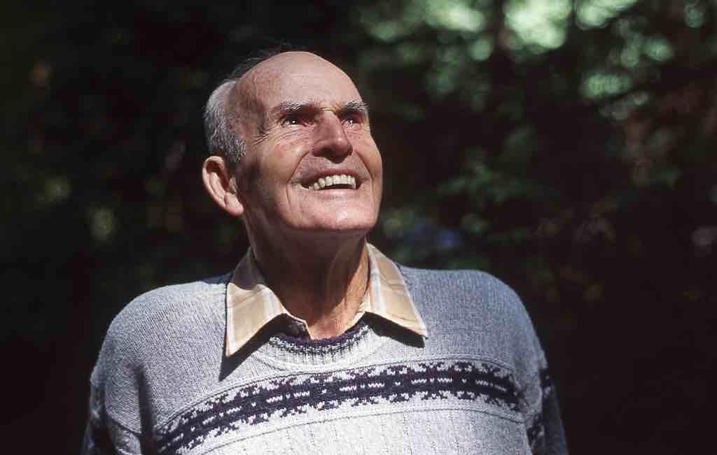 A picture of an elderly man smiling and slightly looking up with trees behind him.