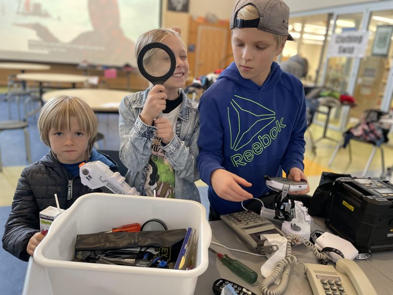 Three young childrent stand at a table that has a bin of tools and other electronics on it, such as an old corded landline phone, a remote, calculators and a printer. One of the children (in the middle) holds a magnifying glass up to their face, looking through it and smiling.