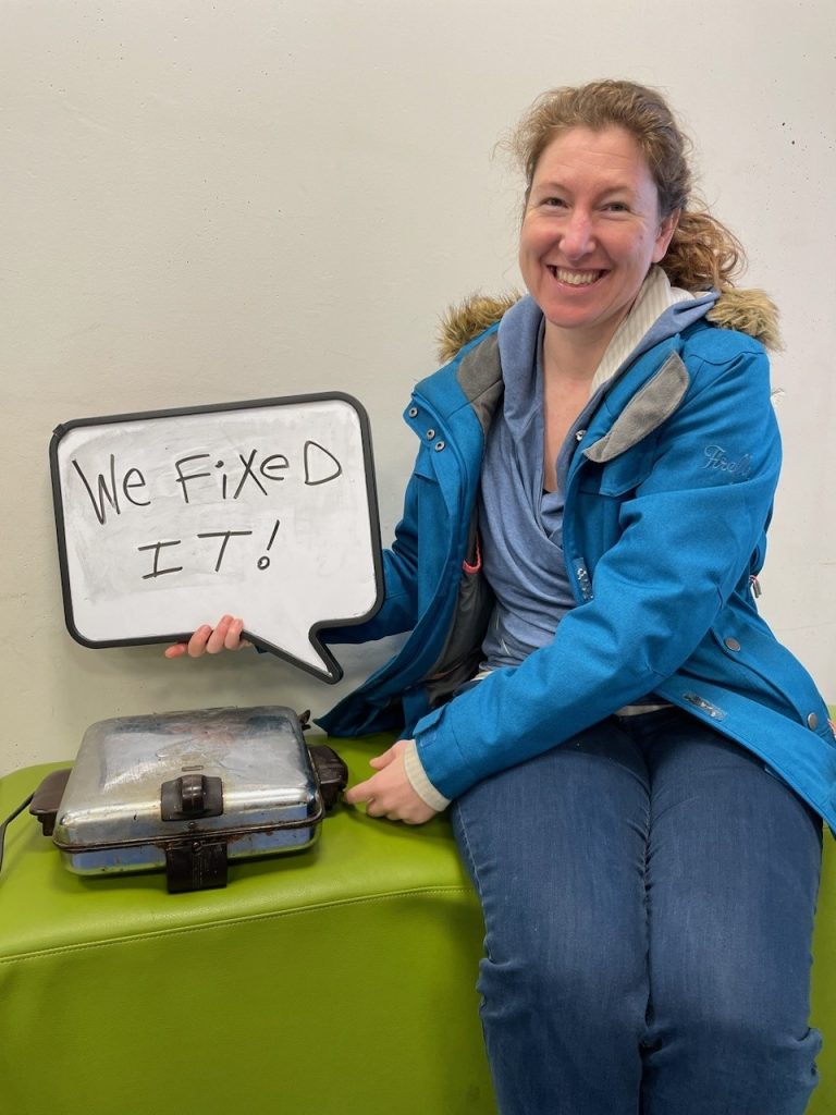 A woman sits next to a griddle that she just had fixed at the Repair Cafe in Courtenay. She holds a sign that says "we fixed it!"