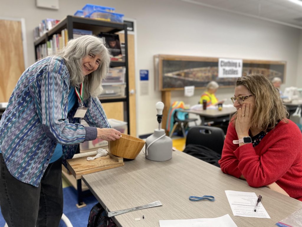 One woman stands next to a table holding a lampshade. Another woman sits at the table with the lamp base in front of her waiting to be repaired.