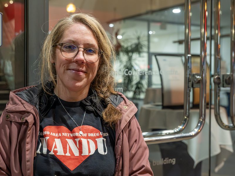 Photo of Sarah Lovegrove standing outside the doors to the Vancouver Island Conference Centre where Nanaimo City Council meets wearing a Nanaimo Area Network of Drug users shirt,