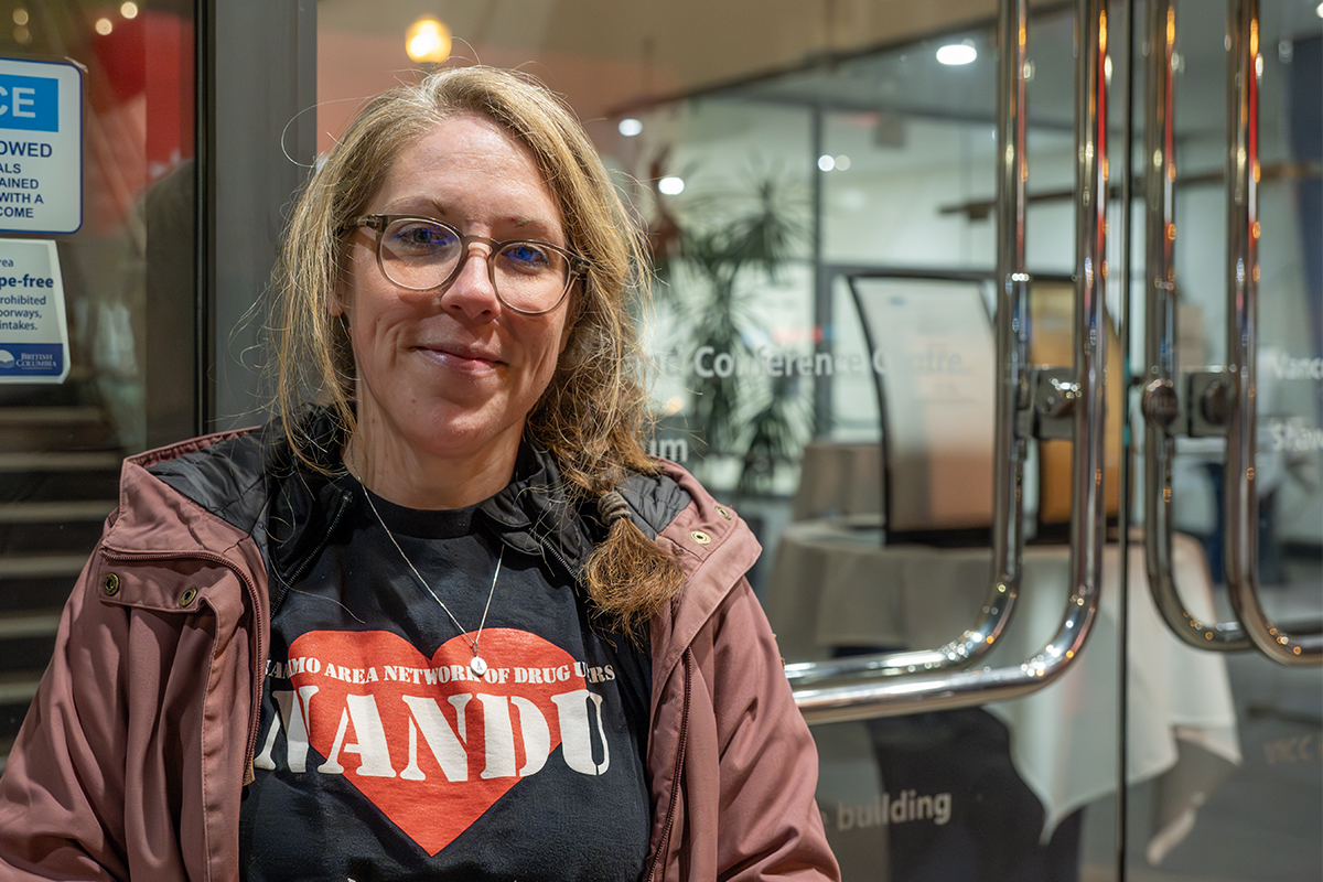 Photo of Sarah Lovegrove standing outside the doors to the Vancouver Island Conference Centre where Nanaimo City Council meets wearing a Nanaimo Area Network of Drug users shirt,