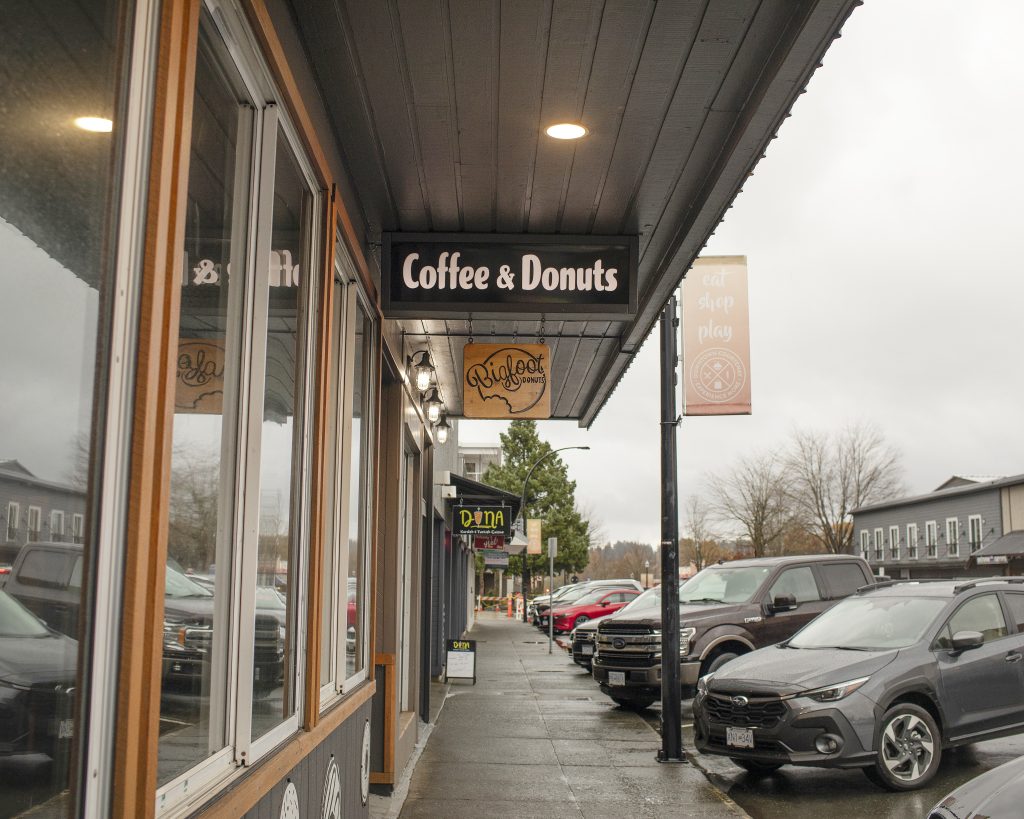 A view of storefronts on the left and cars parked on the right. A sign hanging from an awning reads "Coffee and Donuts" with another sign that says "Bigfoot Donuts" slightly behind it.