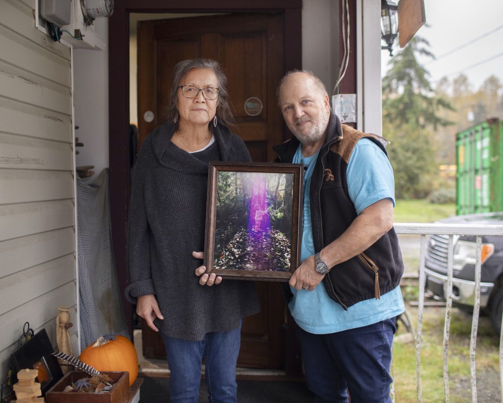 Josephine and Andrew, parents of Kai, stand next to each other holding a framed photo of Kai as a toddler. They are standing in front of a door looking at the camera.