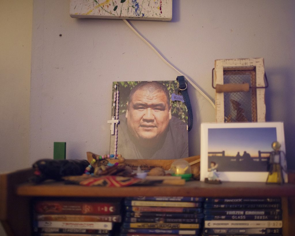 A photograph of a man sits on top of a bookshelf with a rosary with a crucifix hanging from one corner of the photo and what appears to be a dog collar with the name Kelso on the tag on another corner of the photo.