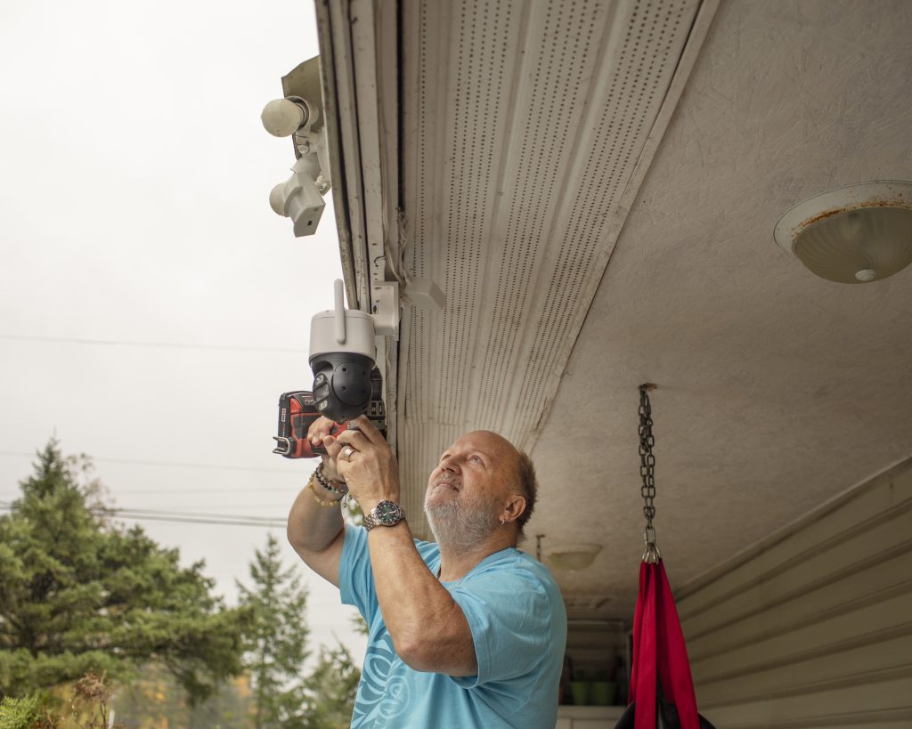 A man, Kai's foster father, installing a security camera to the awning of his home.