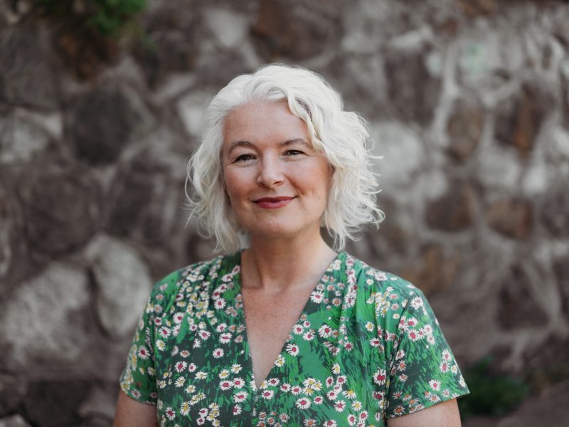 A photo of writer Zena Sharman standing infront of a stone wall.