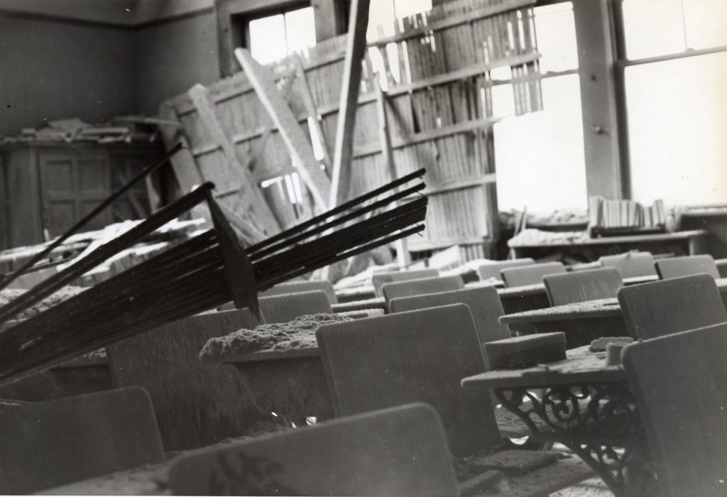 Broken school chairs and tables shown in black and white, from damage to the inside of an elementary school circa 1946