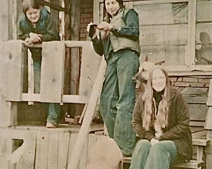 An old photo of three people sitting, standing and slouching in front of a wooden home.