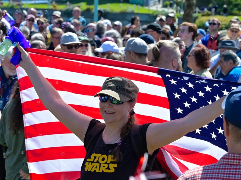 A woman wearing a Stop Wars shirt holds an American Flag and a toy bubble gun at the Nanaimo Infusion in Maffeo Sutton Park on April 25, 2026.