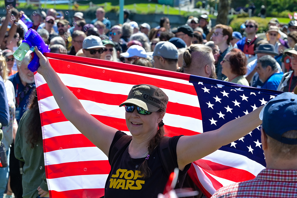 A woman wearing a Stop Wars shirt holds an American Flag and a toy bubble gun at the Nanaimo Infusion in Maffeo Sutton Park on April 25, 2026.