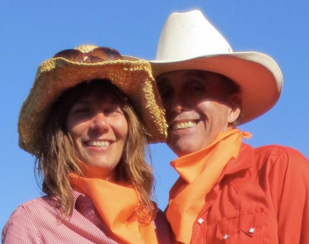 A photo of Vig Schulman on the right smiling beside his wife, Melissa Roeske. They're both wearing cowboy hats, red-orange shirts and orange bandanas around their necks.