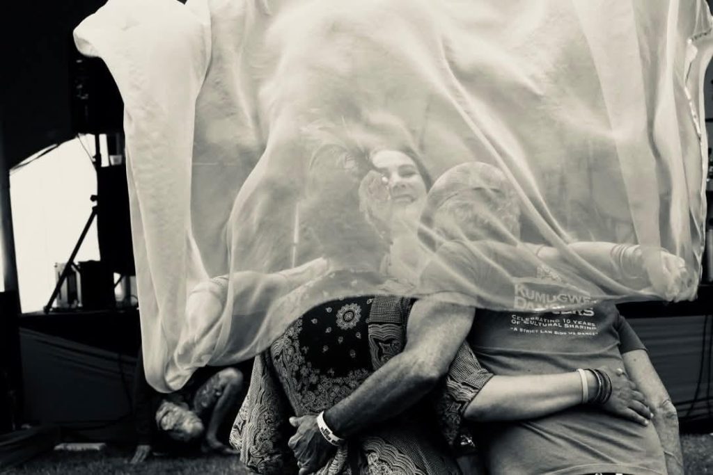 A artistic black and white photo that shows the backs of Vig Schulman and his wife, Melissa, with Vig's arm around her waist. In front of them and facing the camera is smiling Cathy Stoyko who is dancing with some sort of sheer veil she is draping over them.