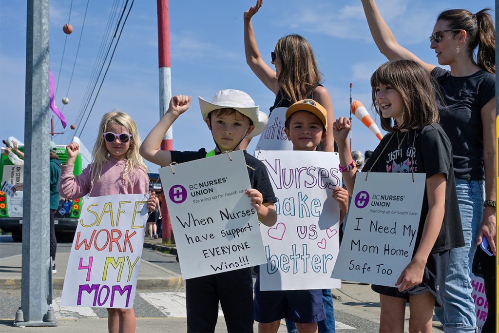 Children ask passing motorists to honk in support of striking nurses during a picket at the Nanaimo Regional General Hospital on July 13, 2026. From left to right: Allie, Sam, Miles and Kenney with their mothers Brittany McKay and Amy Sullivan who work as nurses in Nanaimo.