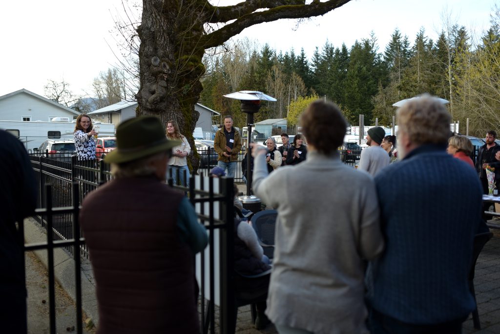 A group of people blurry in the foreground, with people in the background who are in focus. They are gathered outdoors on a patio and appear to be celebrating.