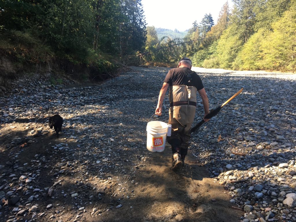A man walks on a dry riverbed with a pail in one hand and a net in the other.