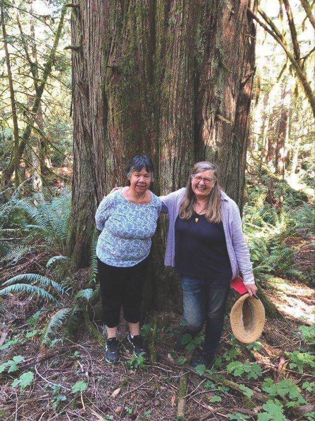 Geraldine Manson and Nancy Turner stand side-by-side in the forest in front of a very big tree.
