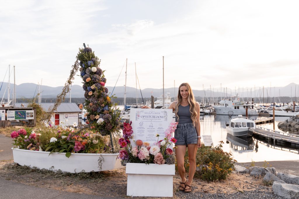 A woman stands next to a dinghy that has been covered in flowers as an installation.