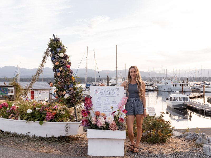 A woman stands next to a dinghy that has been covered in flowers as an installation.