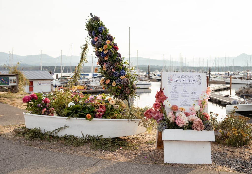 A dinghy on dry land, with the Comox marina behind. The dinghy has been turned into a flower installation