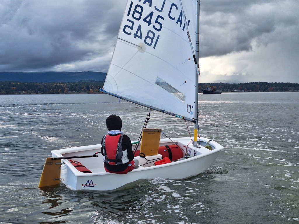 A person is seen sitting on a dinghy. They are turned away from the camera facing the water.