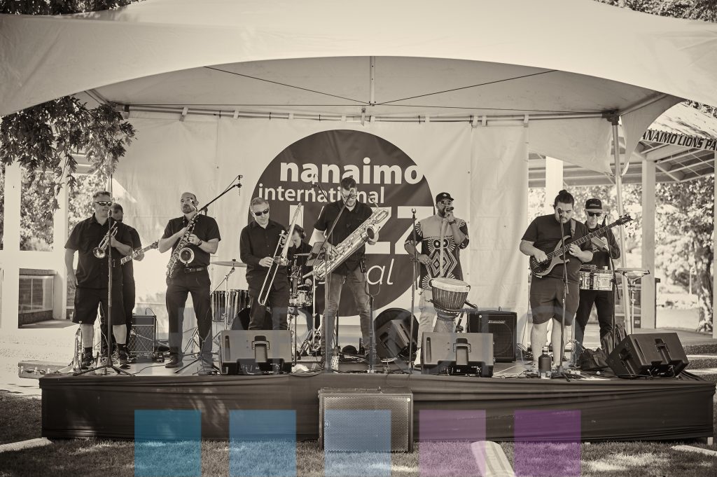 A 9-piece band with various brass instruments performs on a stage in a black and white photo