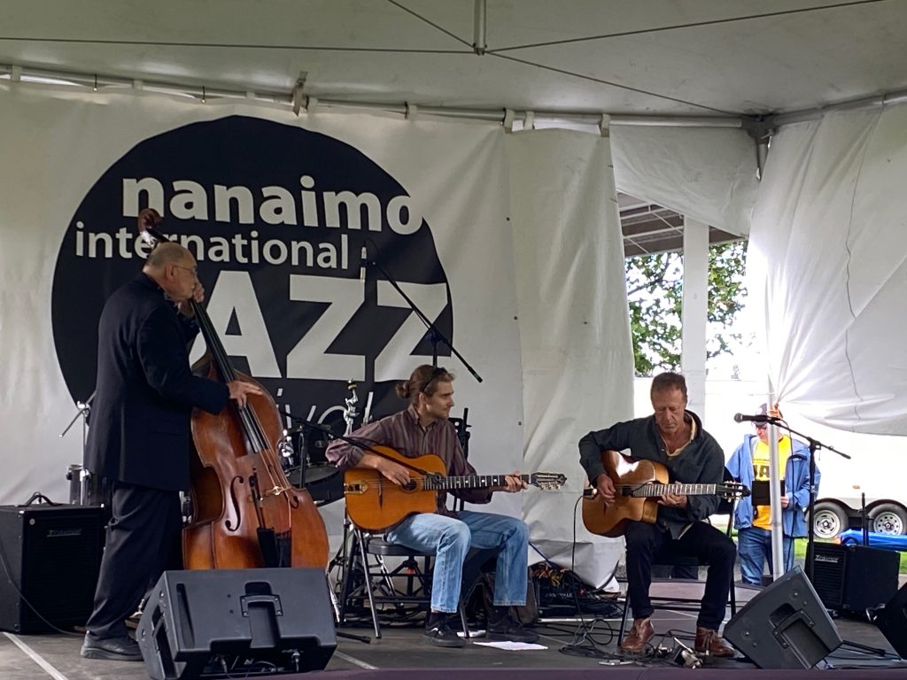 Musicians perform with guitars and an upright bass on a stage with the Nanaimo Jazz Festival logo on the backdrop