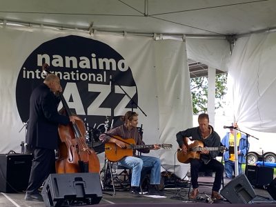Musicians perform with guitars and an upright bass on a stage with the Nanaimo Jazz Festival logo on the backdrop