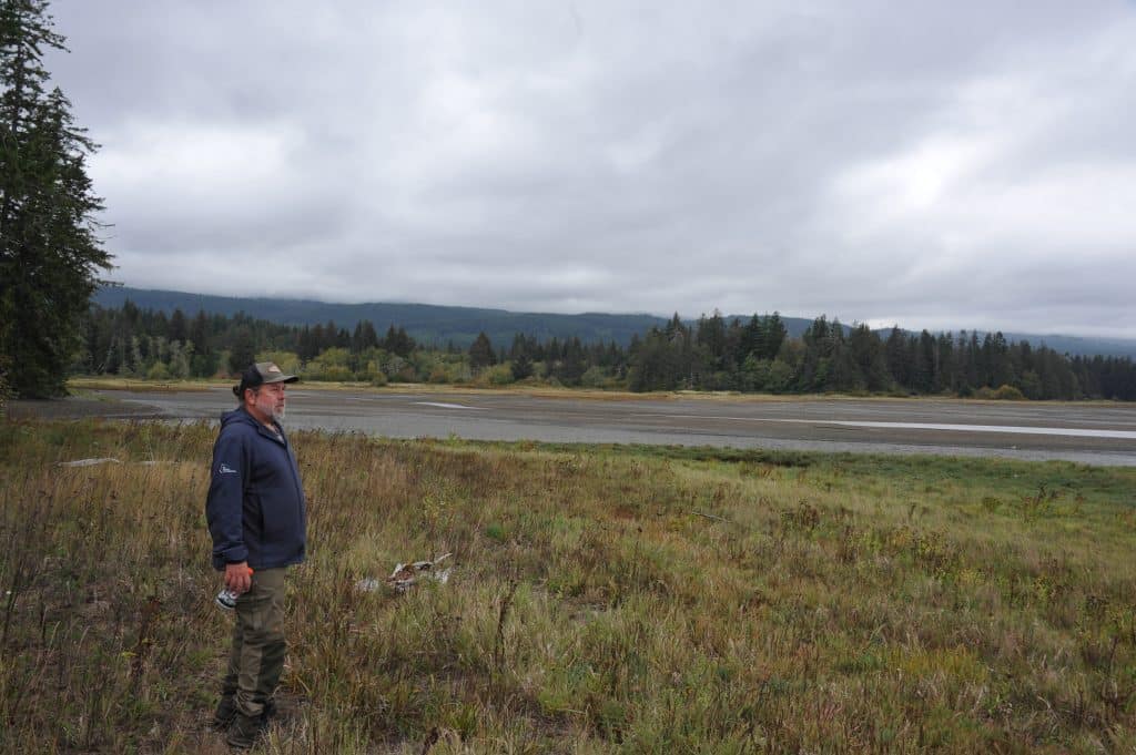 A man stands on the left of the photo looking out towards the right and over a landscape of tall grasses with forest and beach in the background.