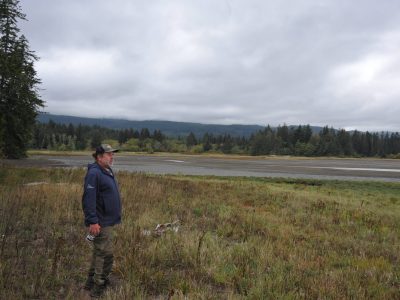 A man stands on the left of the photo looking out towards the right and over a landscape of tall grasses with forest and beach in the background.