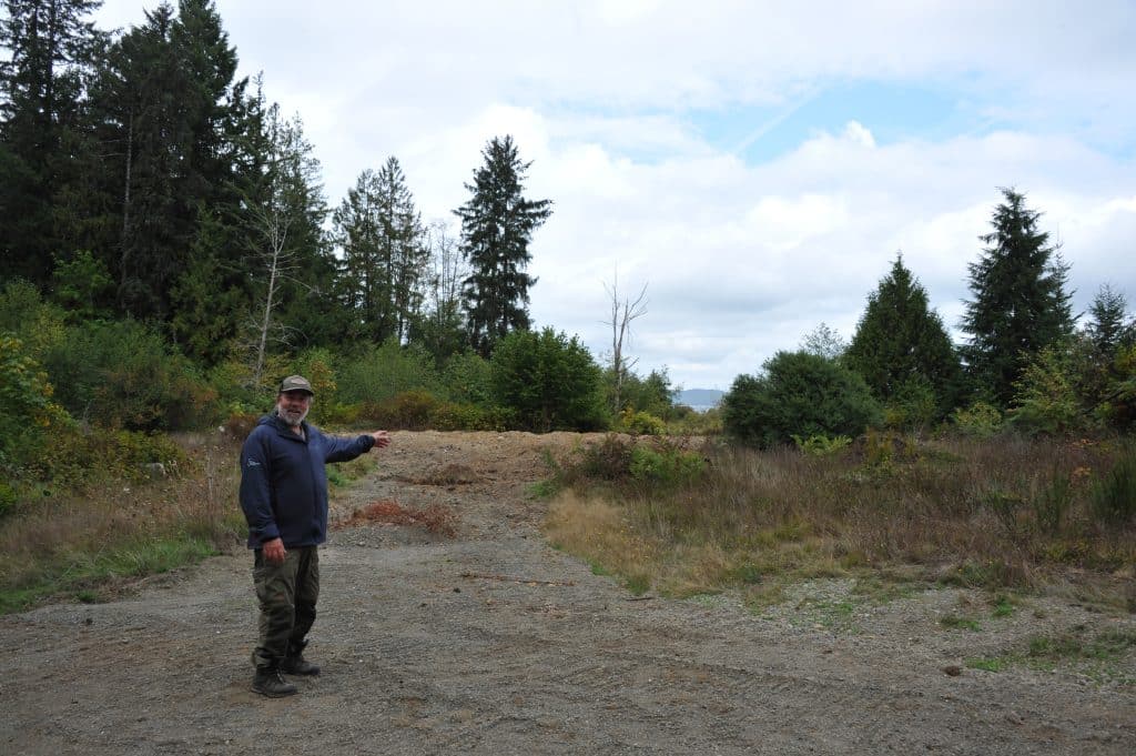 A man stands on a gravel patch and points to a clearing amongst grass and trees that used to be the site of a settler homestead.