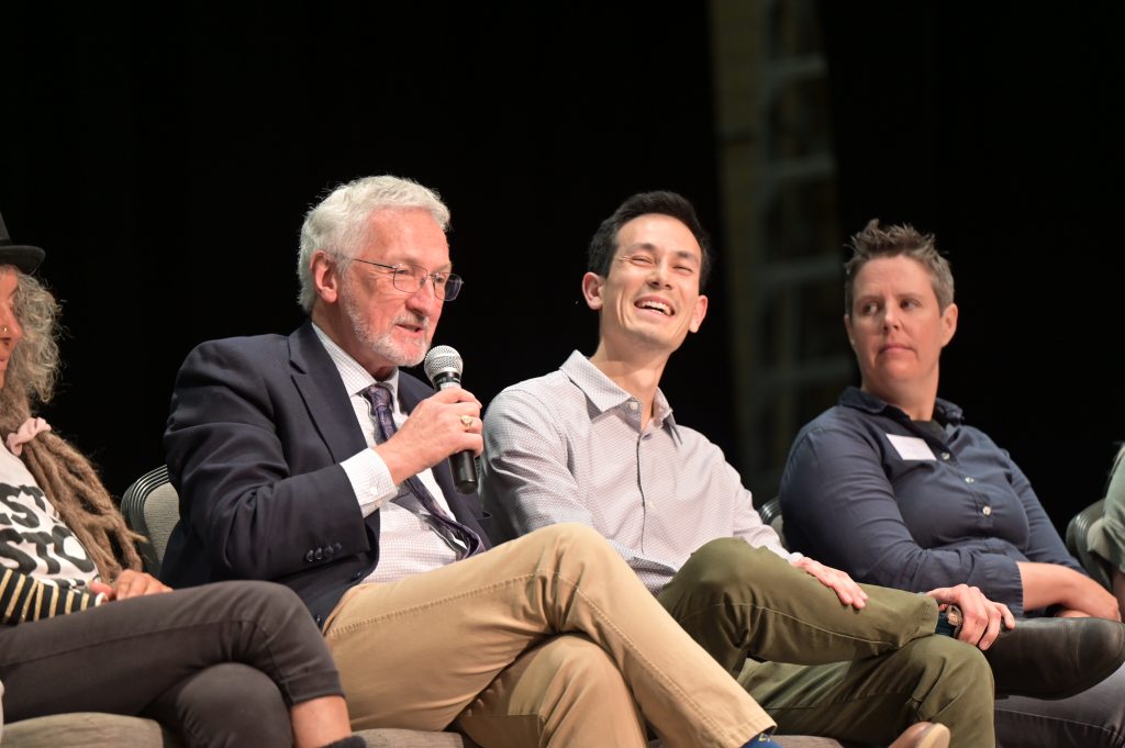 Current Nanaimo Mayor Leonard Krog speaks into a microphone and is seated next to Nanaimo City Council candidate Matthew Miller who is laughing.