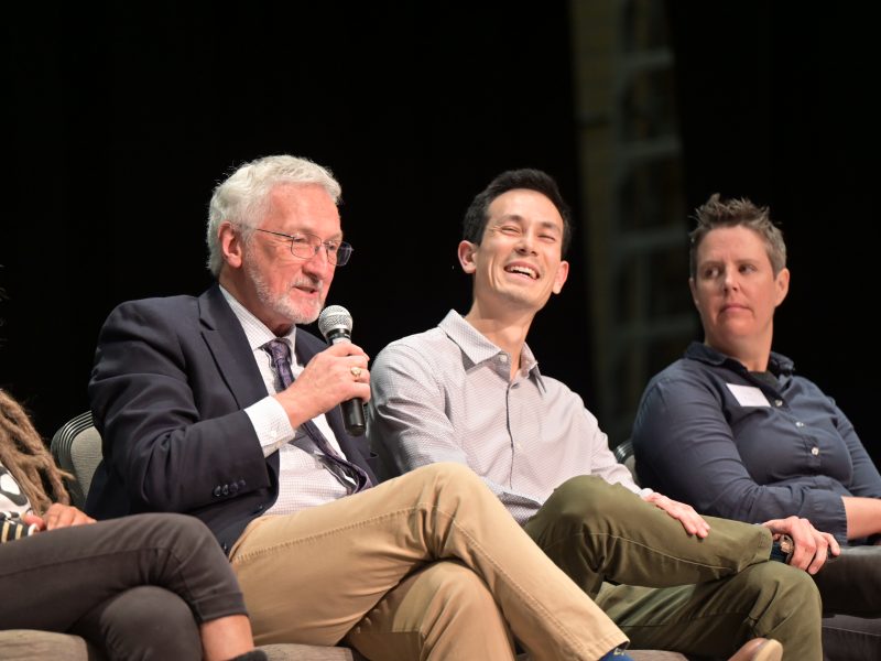 Current Nanaimo Mayor Leonard Krog speaks into a microphone and is seated next to Nanaimo City Council candidate Matthew Miller who is laughing.